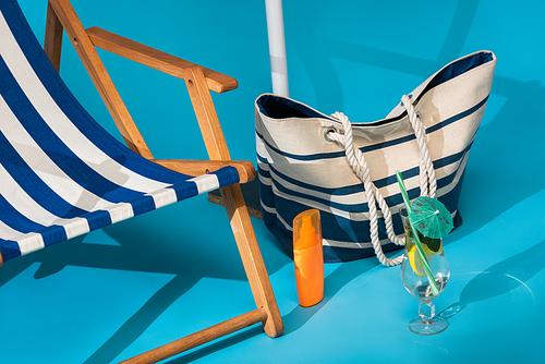 close up view of striped deck chair near sunscreen, beach bag and cocktail on blue background