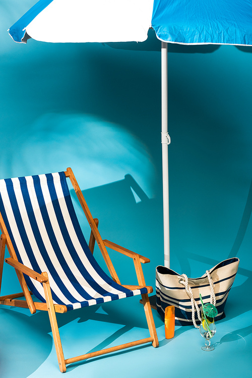 striped deck chair near sunscreen, beach bag and cocktail under umbrella on blue background