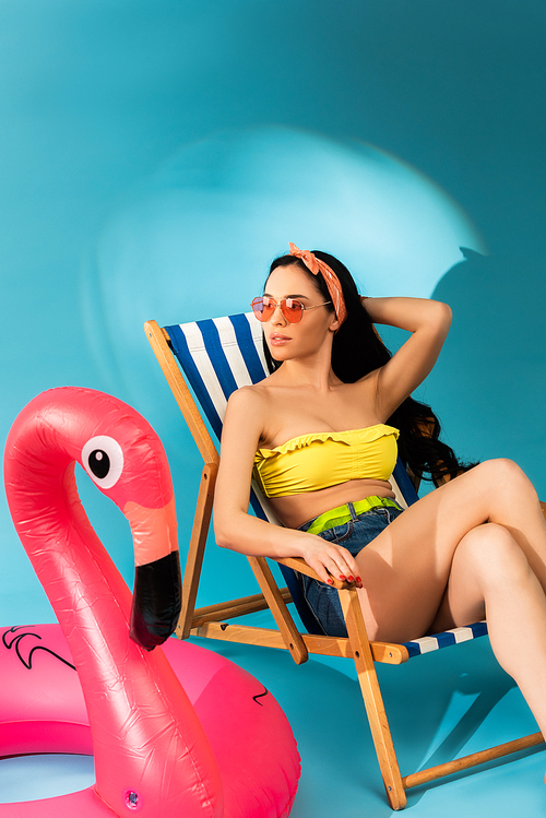 stylish girl sitting in deck chair near inflatable flamingo on blue background