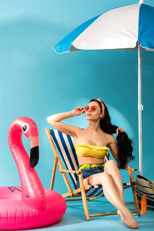 stylish girl sitting in deck chair near inflatable flamingo, beach bag and umbrella on blue background