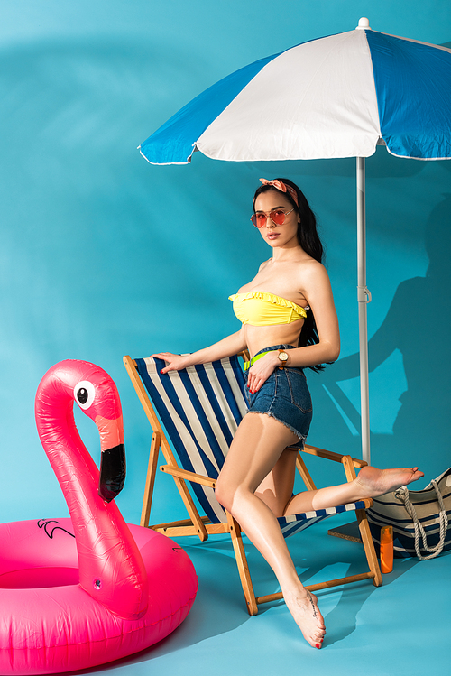stylish girl sitting posing on deck chair near inflatable flamingo, beach bag and umbrella on blue background