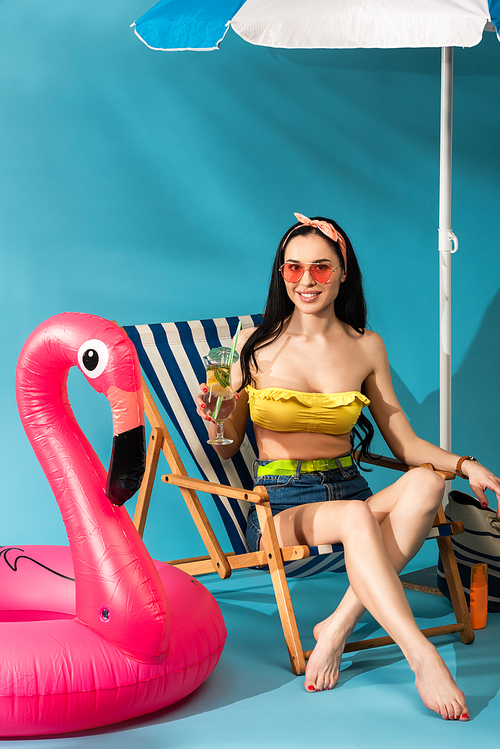 smiling stylish girl sitting in deck chair with cocktail under umbrella near inflatable flamingo on blue background