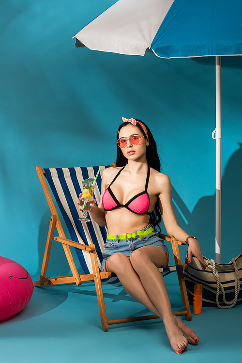 stylish girl sitting in deck chair with cocktail near beach bag and umbrella on blue background