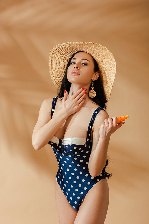 sexy brunette woman in polka dot swimsuit and straw hat applying sunscreen on beige background