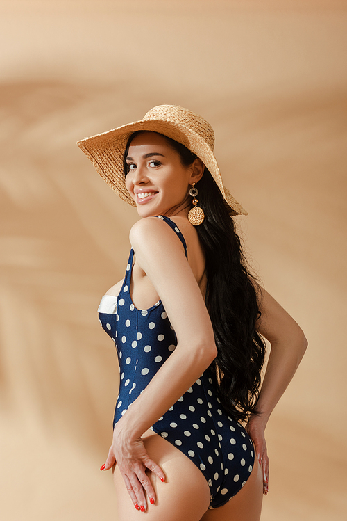 smiling sexy brunette woman in polka dot swimsuit and straw hat posing with hands on buttocks on beige background