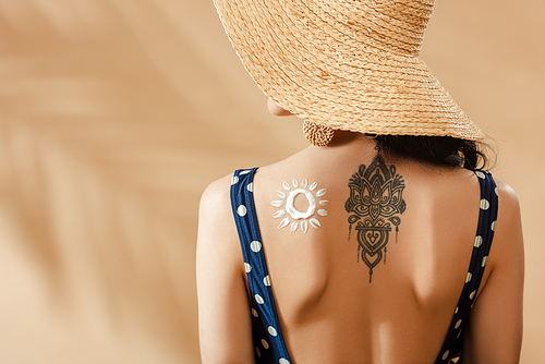 woman in polka dot swimsuit and straw hat with drawn sun and tattoo on back on beige background