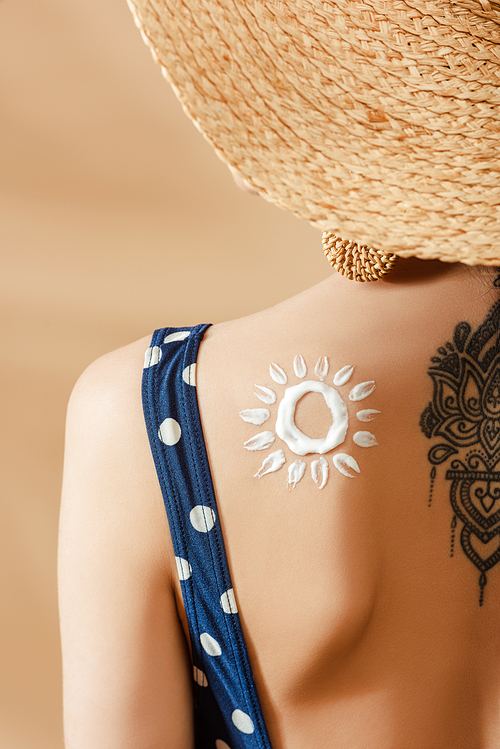 woman in polka dot swimsuit and straw hat with drawn sun and tattoo on back on beige background