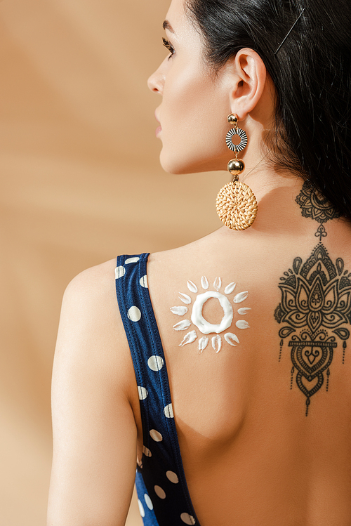 back view of sexy brunette woman in polka dot swimsuit with drawn sun made of sunscreen and tattoo on back on beige background