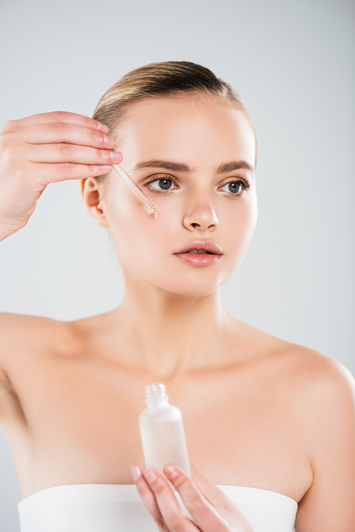 attractive woman holding bottle and pipette while applying serum isolated on grey