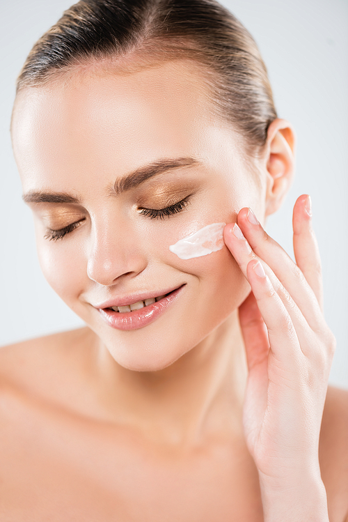 cheerful woman with closed eyes applying face cream isolated on grey