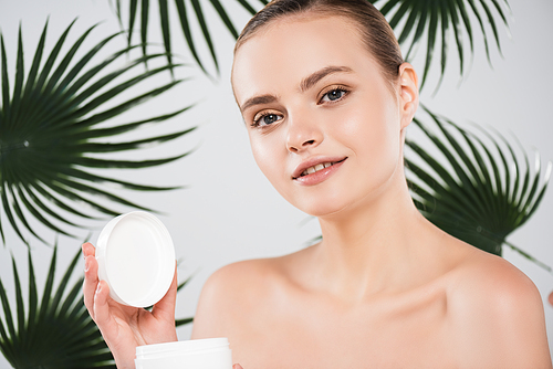 happy woman holding container with face cream near palm leaves on white