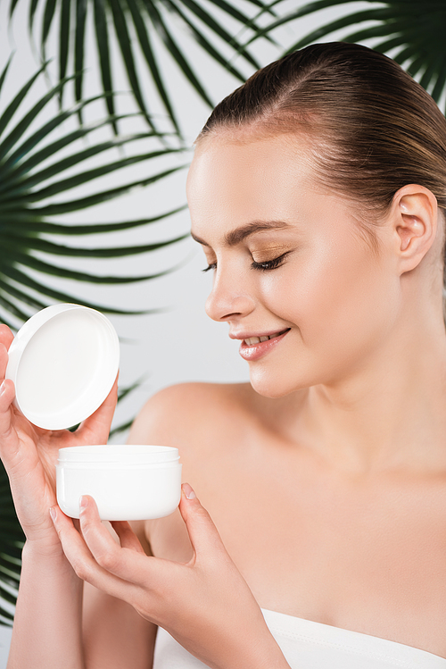 happy woman looking at container with face cream near palm leaves on white