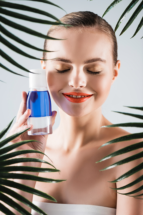 happy woman with makeup holding bottle with toner near palm leaves on white
