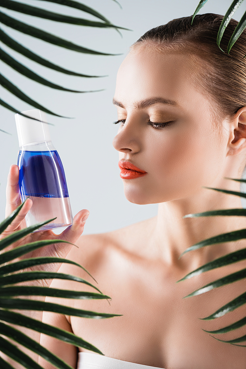 attractive woman with makeup looking at bottle with toner near palm leaves on white