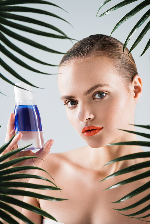 attractive girl with makeup holding bottle with toner near palm leaves on white