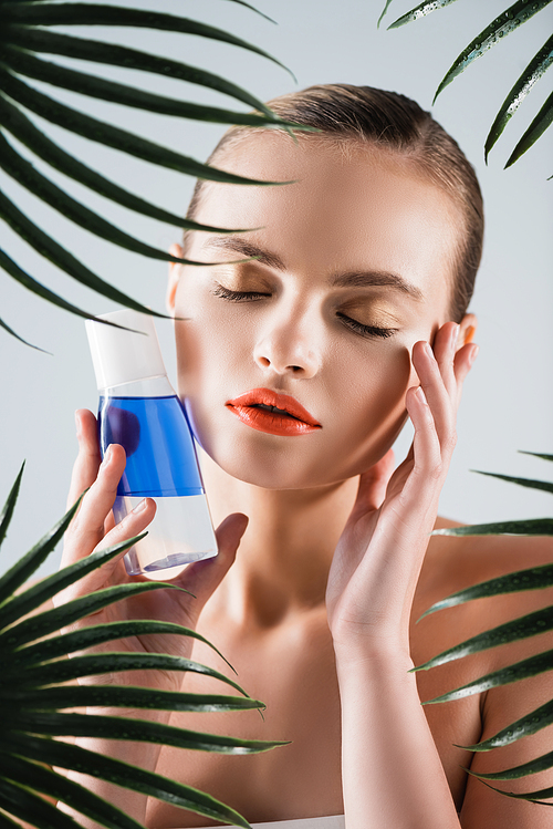 attractive girl with makeup holding bottle with toner and touching face near palm leaves on white