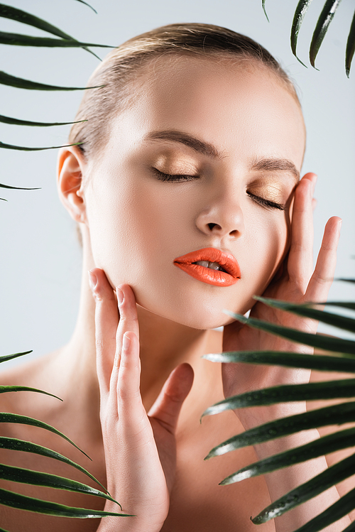 selective focus of attractive girl with makeup touching face near palm leaves on white