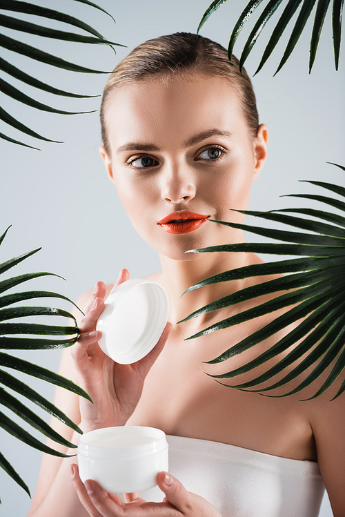attractive girl with makeup holding container with face cream near palm leaves on white