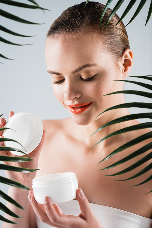happy girl with makeup holding container with face cream near palm leaves on white