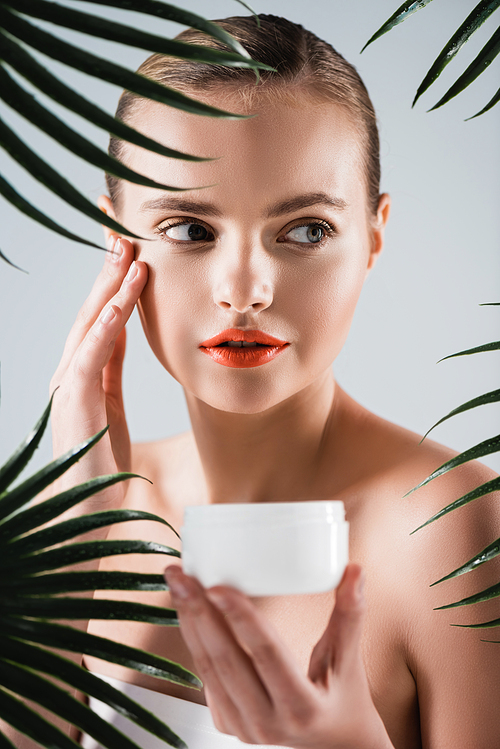 attractive girl with makeup holding container with face cream and touching face near palm leaves on white