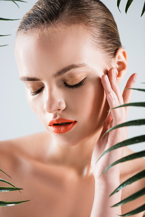 attractive and naked girl with makeup touching face near palm leaves on white