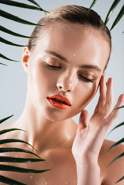 attractive and wet woman with makeup touching face near palm leaves on white