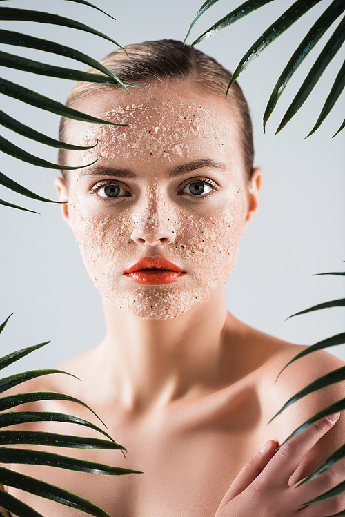 naked woman with makeup and scrub on face  near palm leaves on white
