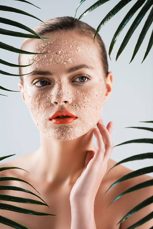 selective focus of naked woman with scrub touching face near palm leaves on white