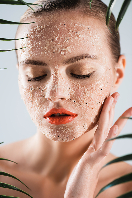 selective focus of naked woman with makeup and scrub touching face near palm leaves on white