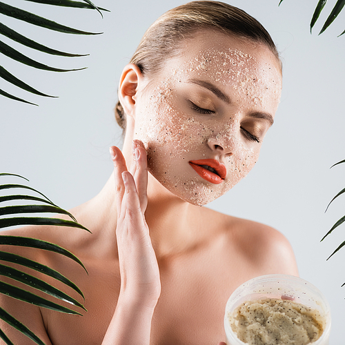 naked woman applying scrub and holding container near palm leaves on white