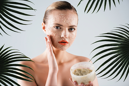 attractive young woman applying scrub and holding container near palm leaves on white