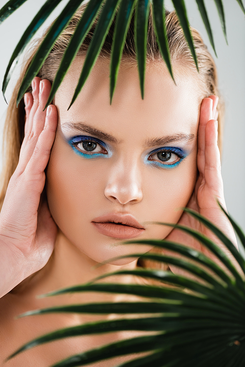 selective focus of beautiful woman with with blue eye shadow touching face near palm leaves on white