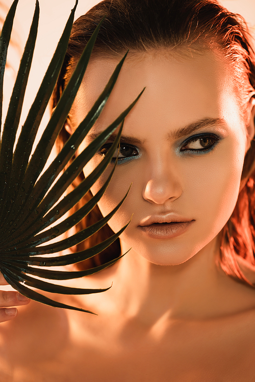 close up of beautiful woman with makeup looking away near palm leaf