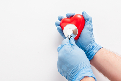 cropped view of doctor in latex gloves examining red heart with stethoscope on white background