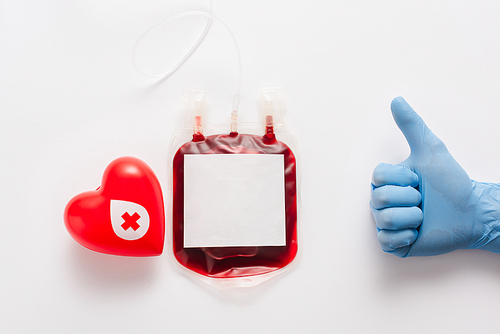 cropped view of doctor showing thumb up near blood package and red heart on white background