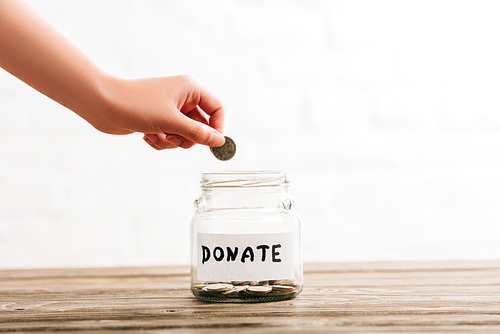 cropped view of woman putting coin in penny jar with donate lettering on wooden surface on white background