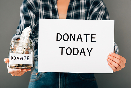 cropped view of woman holding card with donate today lettering and jar with money on grey background