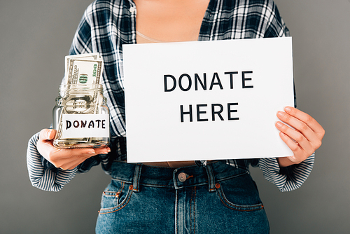 cropped view of woman holding card with donate here lettering and jar with money on grey background
