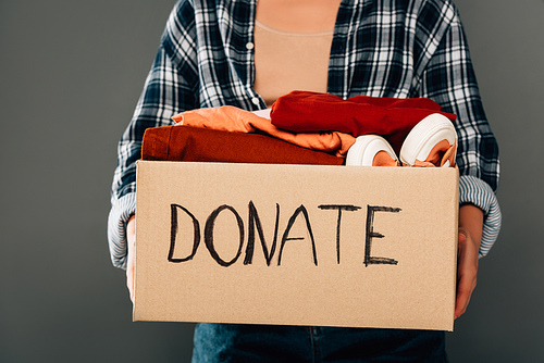 cropped view of woman holding box with donate lettering and clothes on grey background