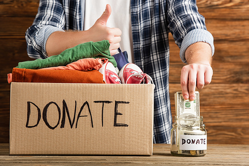 cropped view of man showing thumb up near cardboard box with donated clothes and money on wooden background, charity concept