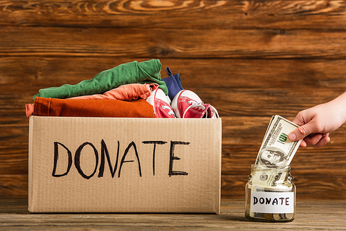 cropped view of man putting money in jar near cardboard box with donate lettering and clothes on wooden background, charity concept