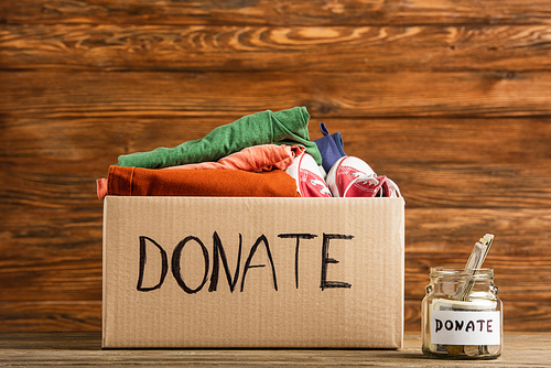 money in jar near cardboard box with donate lettering and clothes on wooden background, charity concept