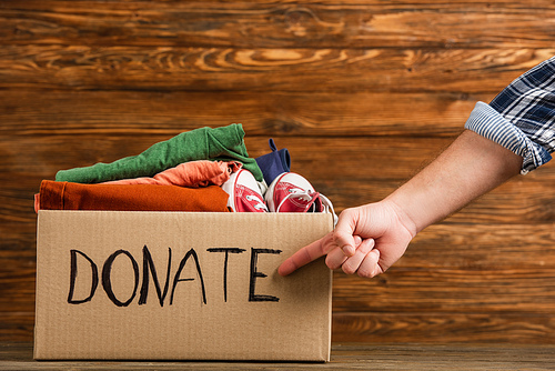 cropped view of man pointing at cardboard box with donate lettering and clothes on wooden background, charity concept