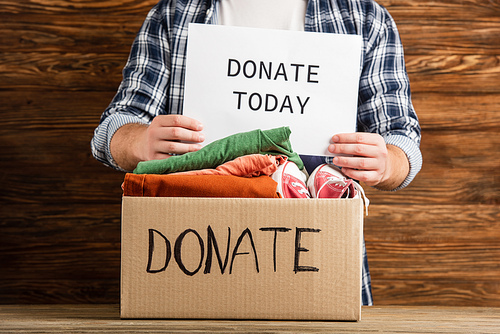 cropped view of man holding donate today card near cardboard box with clothes on wooden background, charity concept