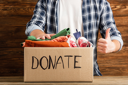 cropped view of man showing thumb up near cardboard box with donate lettering and clothes on wooden background, charity concept