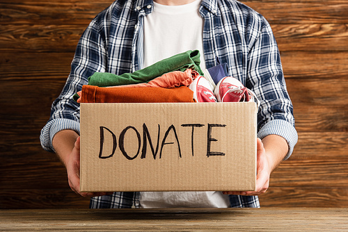 cropped view of man holding cardboard box with donate lettering and clothes on wooden background, charity concept