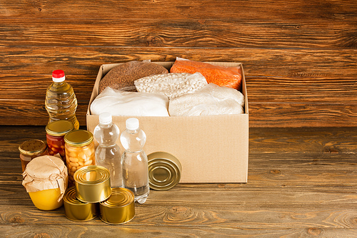 cardboard box with groats near water, oil, canned food and honey on wooden background, charity concept