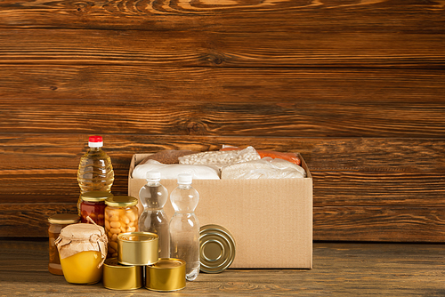 cardboard box with groats near water, oil, canned food and honey on wooden background, charity concept