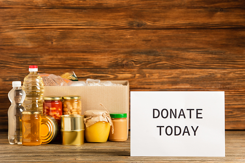 cardboard box with groats near water, oil, canned food, honey and donate today card on wooden background, charity concept