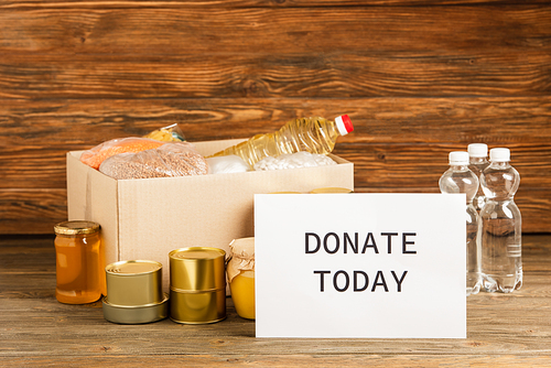 cardboard box with groats near water, oil, canned food, honey and donate today card on wooden background, charity concept